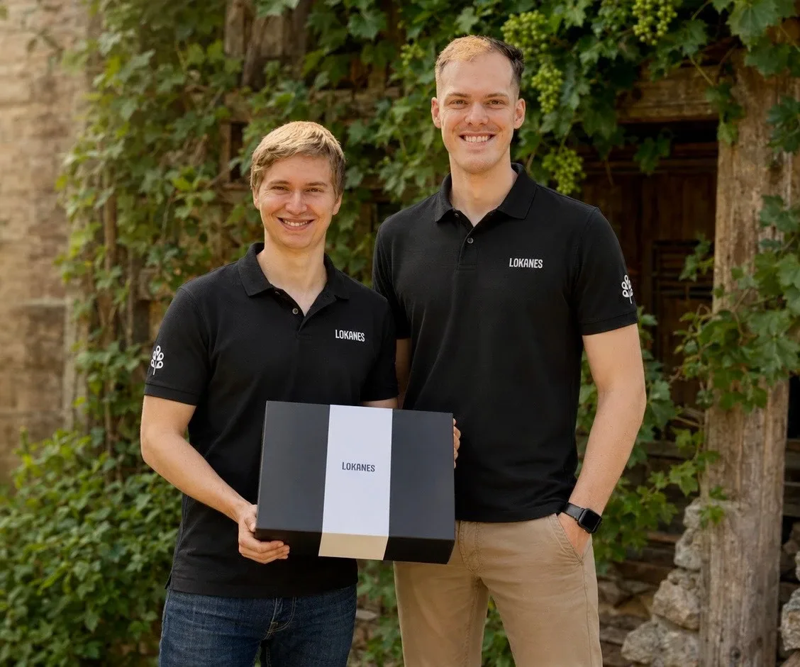 Die beiden Gründer in schwarzen Poloshirts mit einer LOKANES-Geschenkbox, im Freien vor Weinlaub.